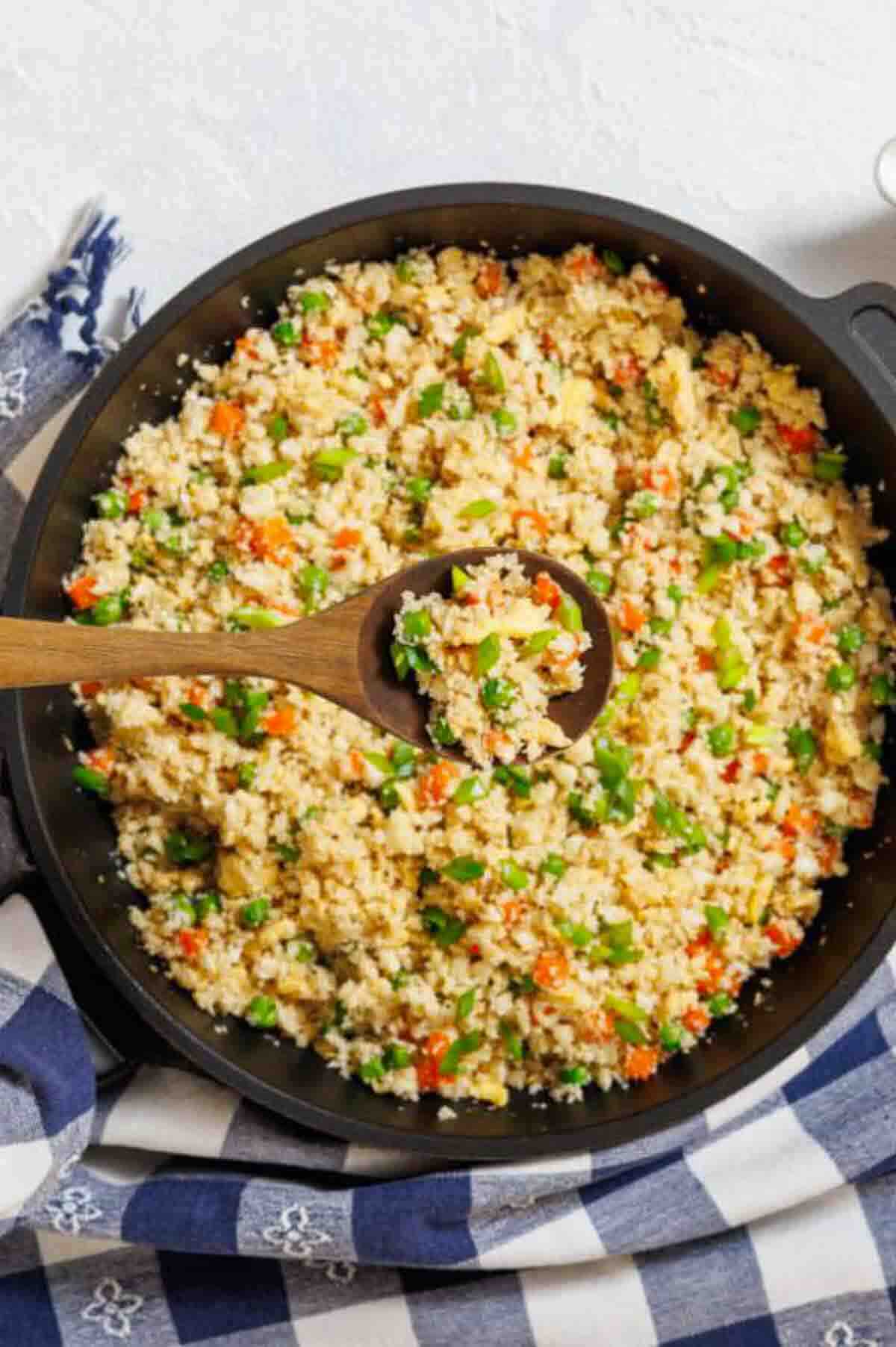 A wooden spoon rests in a skillet filled with cauliflower fried rice mixed with chopped vegetables like carrots and green onions.