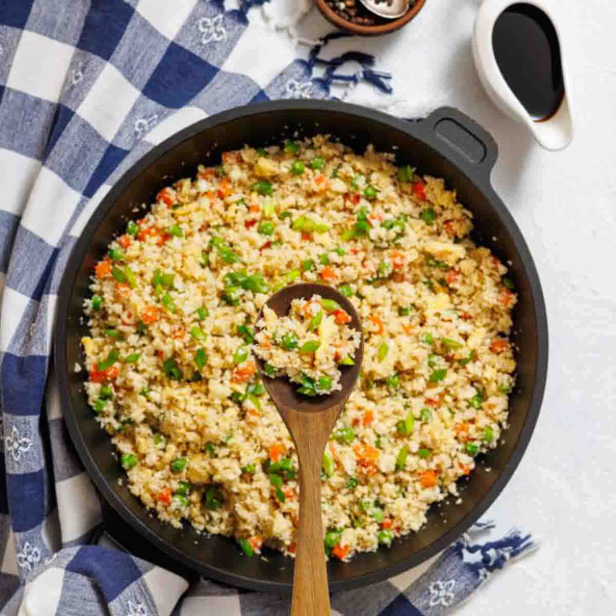 Overhead view of a skillet filled with vegetable fried rice, with a wooden spoon placed on top. A small pitcher of soy sauce is nearby on a checkered cloth.