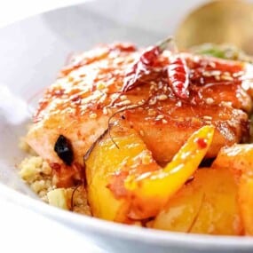 A close-up of a plated dish featuring glazed salmon, yellow bell peppers, dried red chili peppers, and a sprinkle of sesame seeds on a bed of couscous.