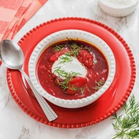 A bowl of borscht topped with sour cream and fresh dill, placed on a red plate with a spoon on the side.
