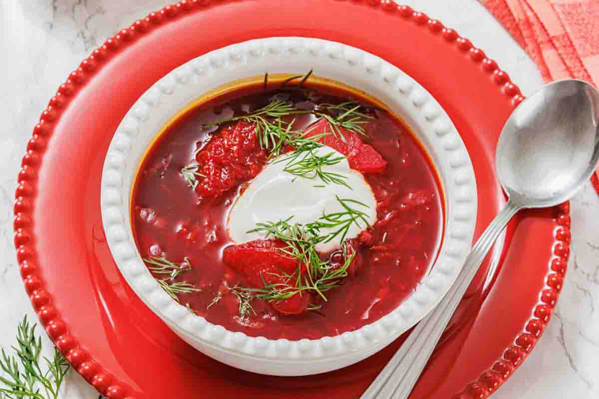 A bowl of borscht topped with a dollop of sour cream and fresh dill, served on a red plate with a spoon beside it.