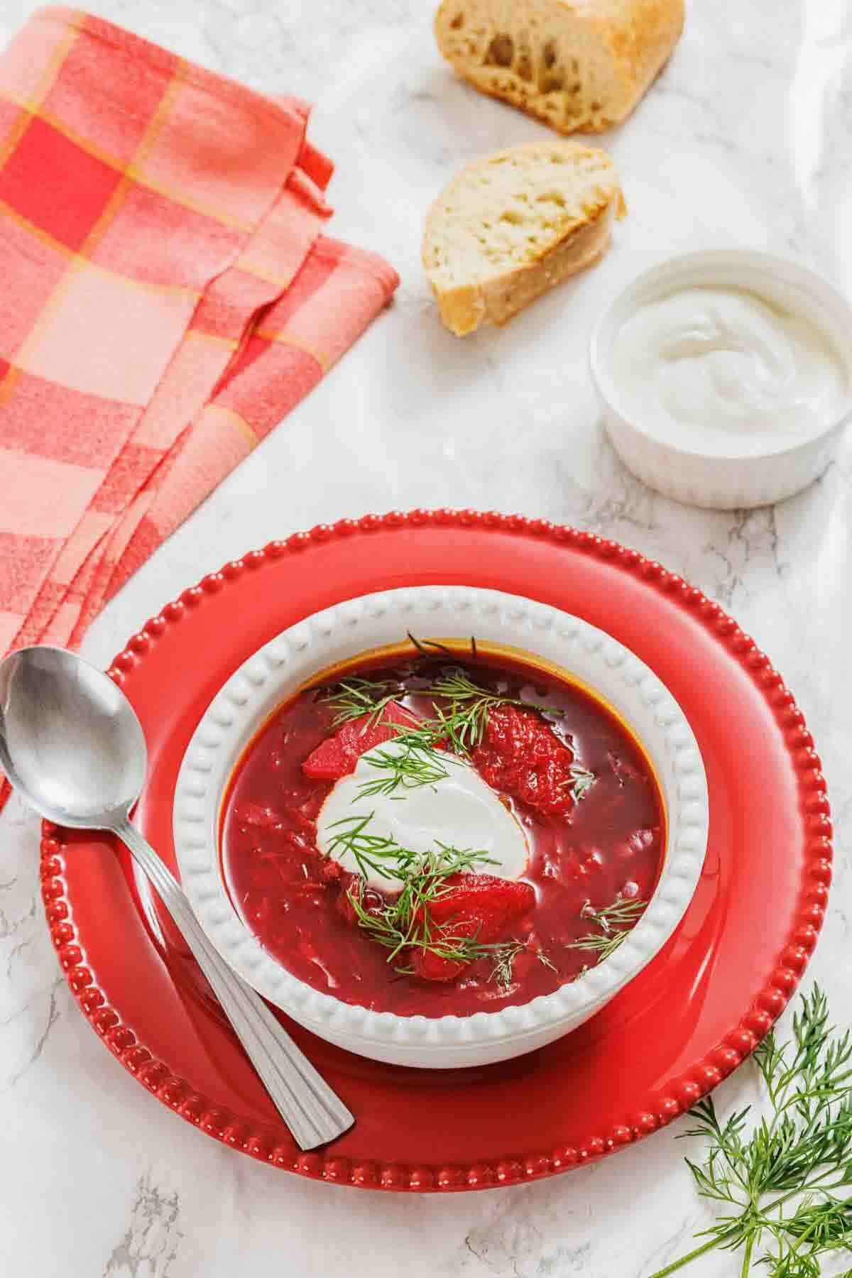 A bowl of red beet soup garnished with dill and a dollop of sour cream, served with a spoon on a red plate. Bread, sour cream, and a red napkin are nearby on a marble surface.