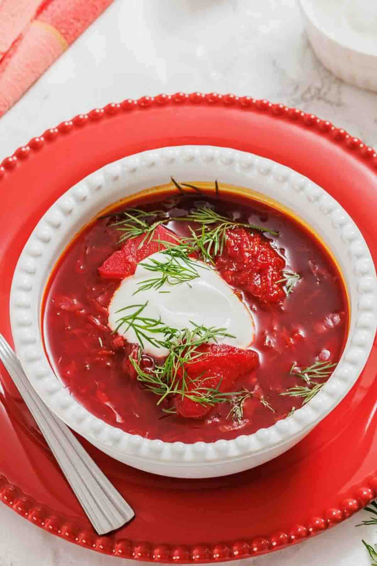 A bowl of borscht soup garnished with fresh dill and a dollop of sour cream, served on a red plate with a spoon.
