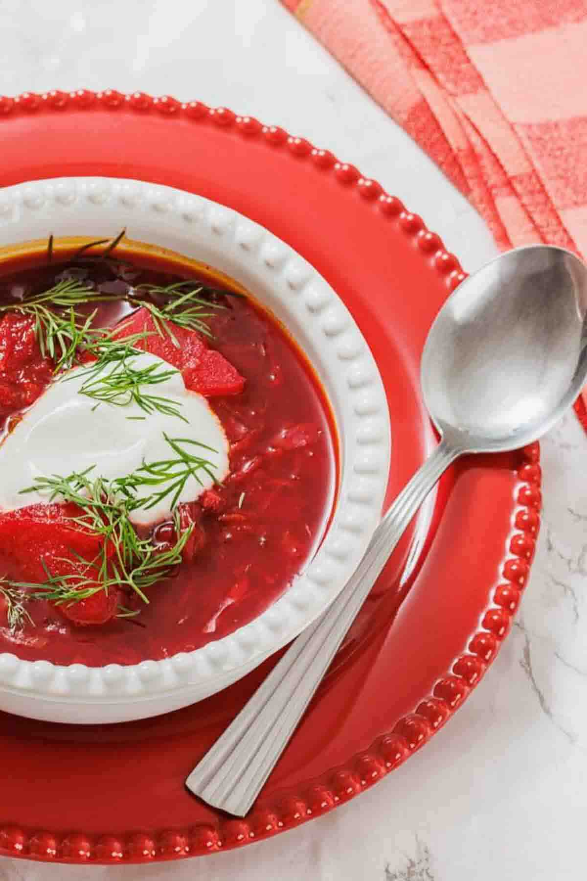 A bowl of borscht topped with sour cream and fresh dill, served on a red plate with a spoon beside it.