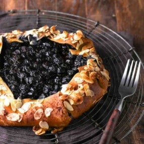 A rustic blueberry galette topped with sliced almonds on a wire cooling rack next to a fork.