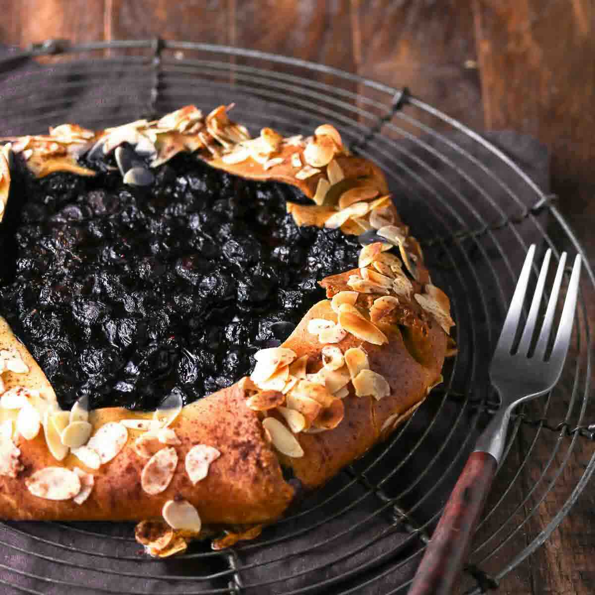 A rustic blueberry galette topped with sliced almonds on a wire cooling rack next to a fork.