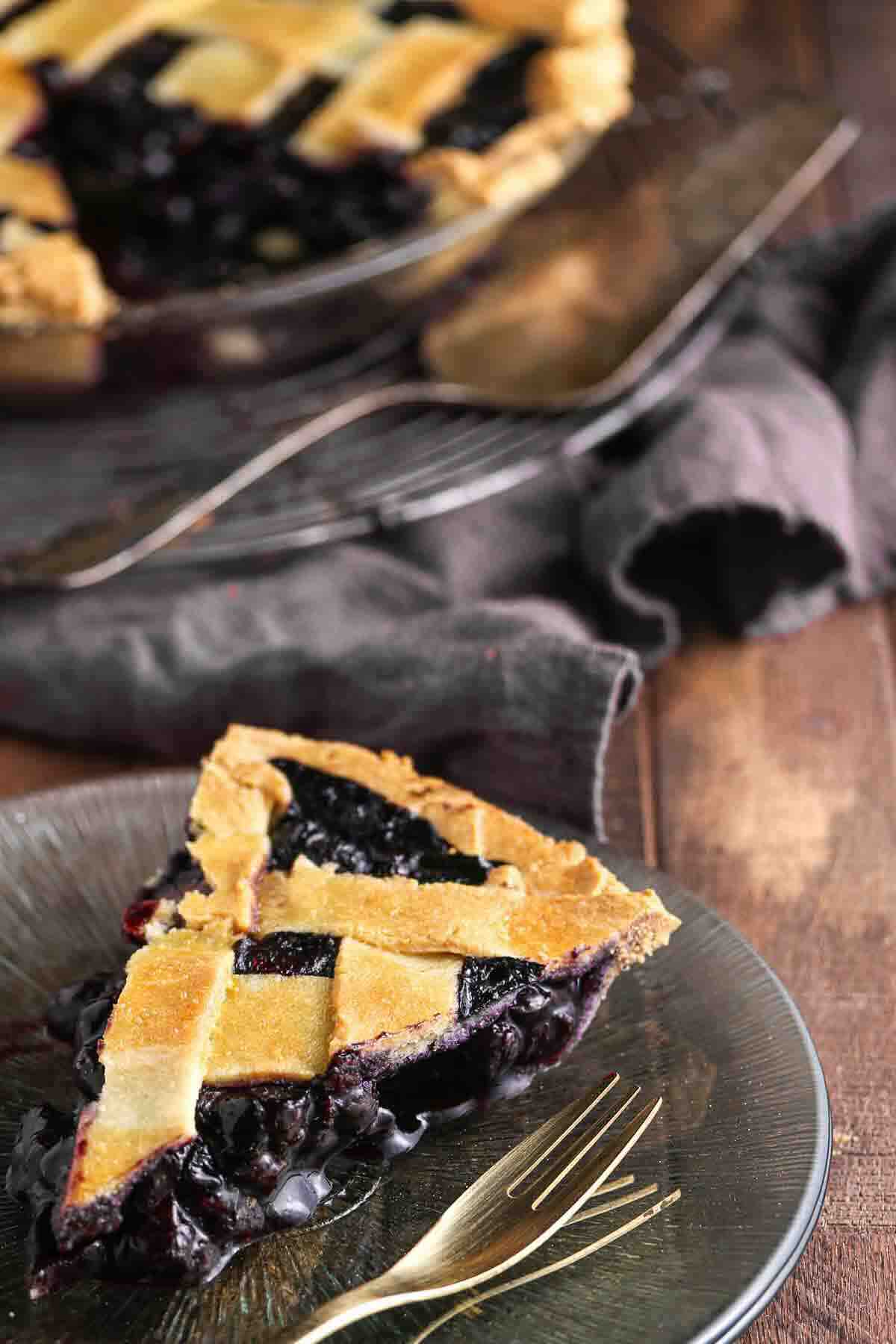 A slice of blueberry pie with a lattice crust sits on a glass plate with a gold fork; the rest of the pie is in the background on a wooden surface.