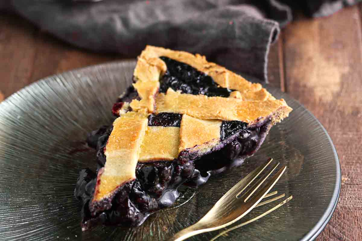 A slice of blueberry pie with a lattice crust sits on a glass plate next to a gold fork.