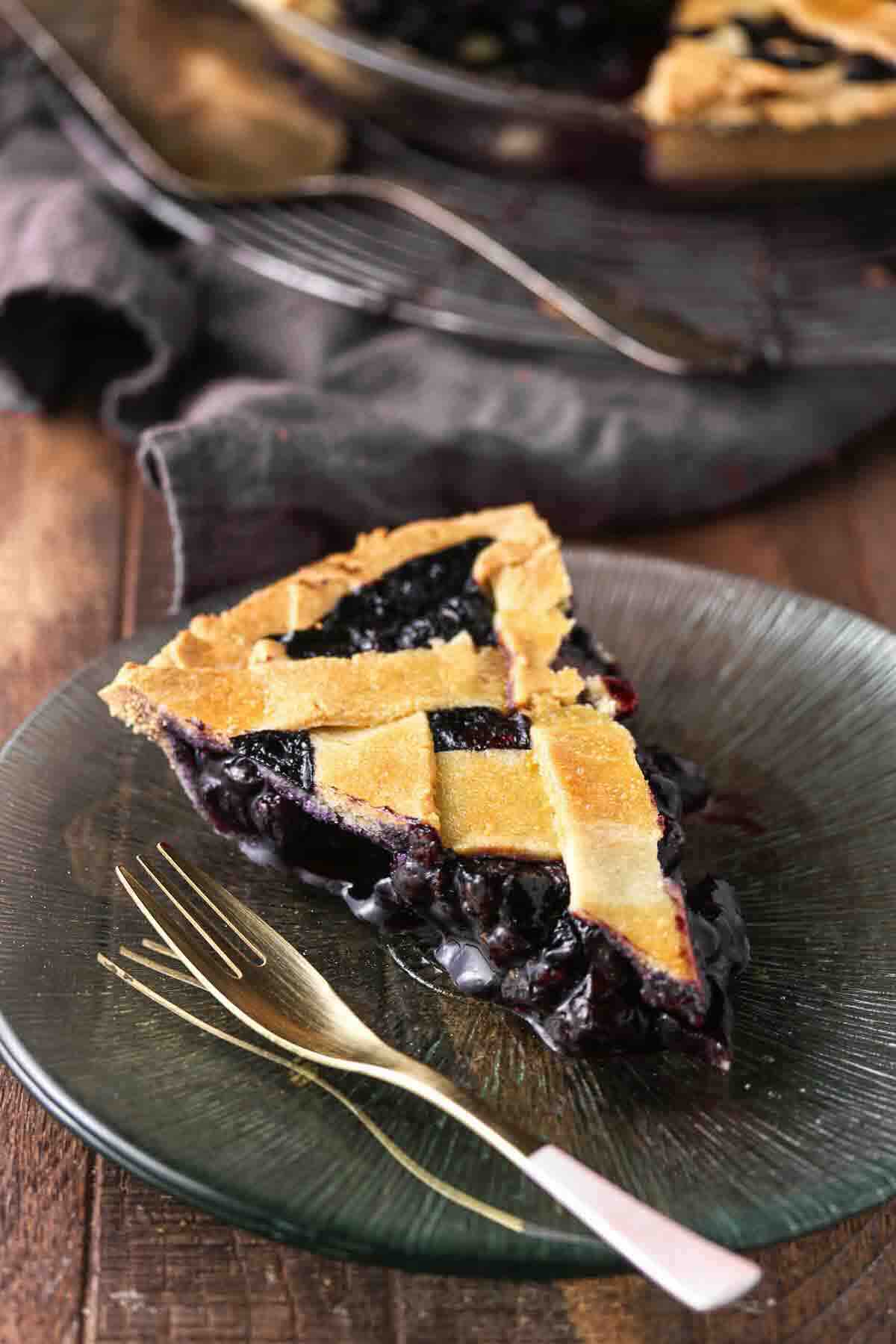 A slice of blueberry pie with a lattice crust on a glass plate, next to a fork, set on a wooden table.