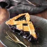 A slice of blueberry pie with a lattice crust sits on a dark glass plate next to a gold fork.