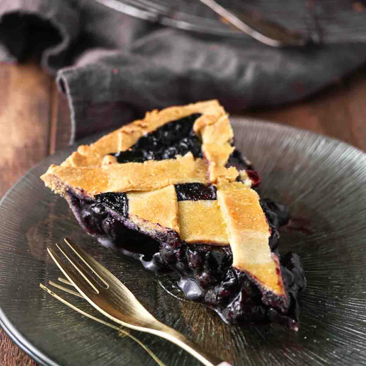 A slice of blueberry pie with a lattice crust sits on a dark glass plate next to a gold fork.