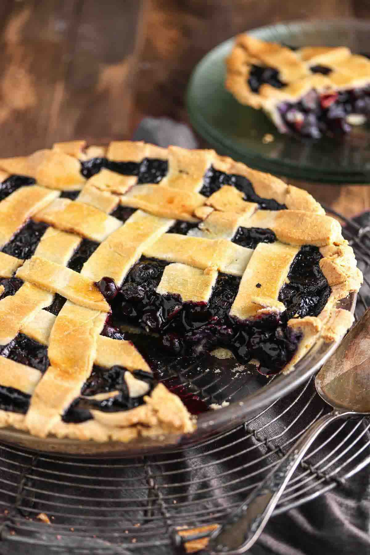A lattice-topped blueberry pie in a glass dish with a slice missing, and the slice placed on a green plate in the background.