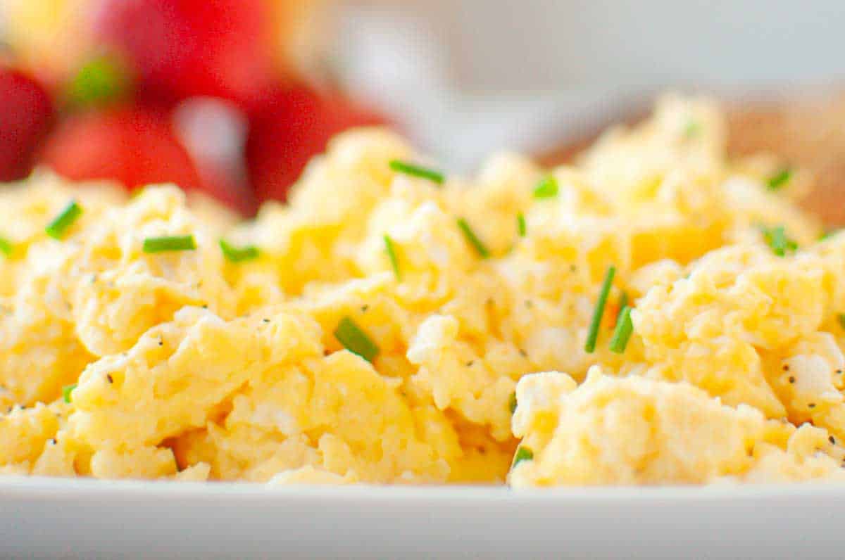 Close-up of scrambled eggs garnished with chopped chives on a white plate, with blurred fruit and toast in the background.