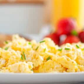 A plate of scrambled eggs garnished with chopped chives, with blurred strawberries and orange juice in the background.