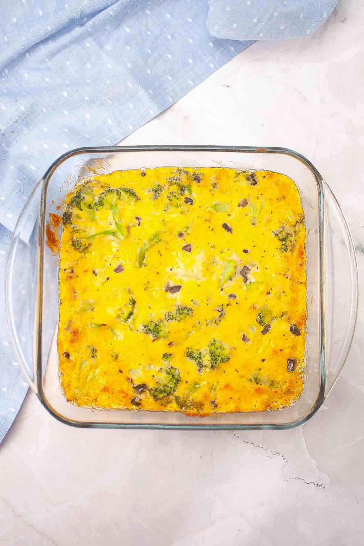 A baked egg casserole with broccoli and diced red onion in a square glass dish, placed on a white surface with a blue cloth nearby.