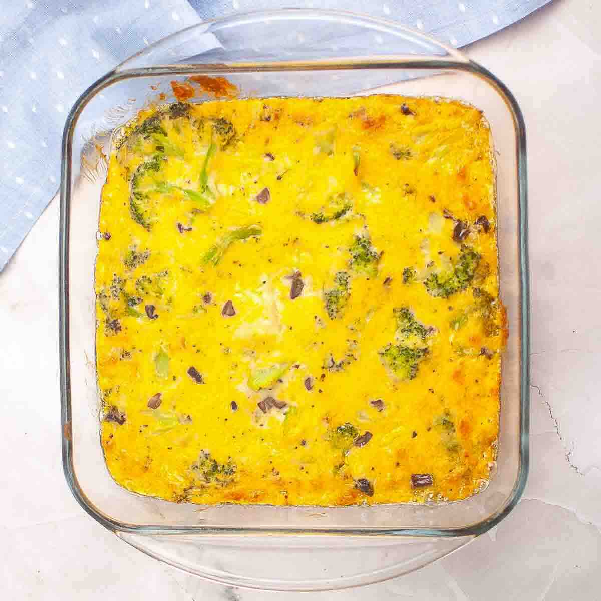 A square glass baking dish filled with a baked egg casserole containing broccoli and bits of purple onion, set on a light surface.