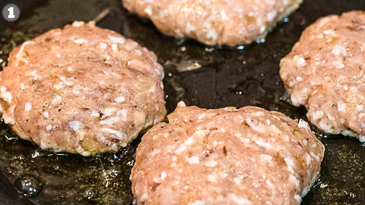 Four raw burger patties with visible bits of onion and seasoning are cooking in oil on a hot skillet.