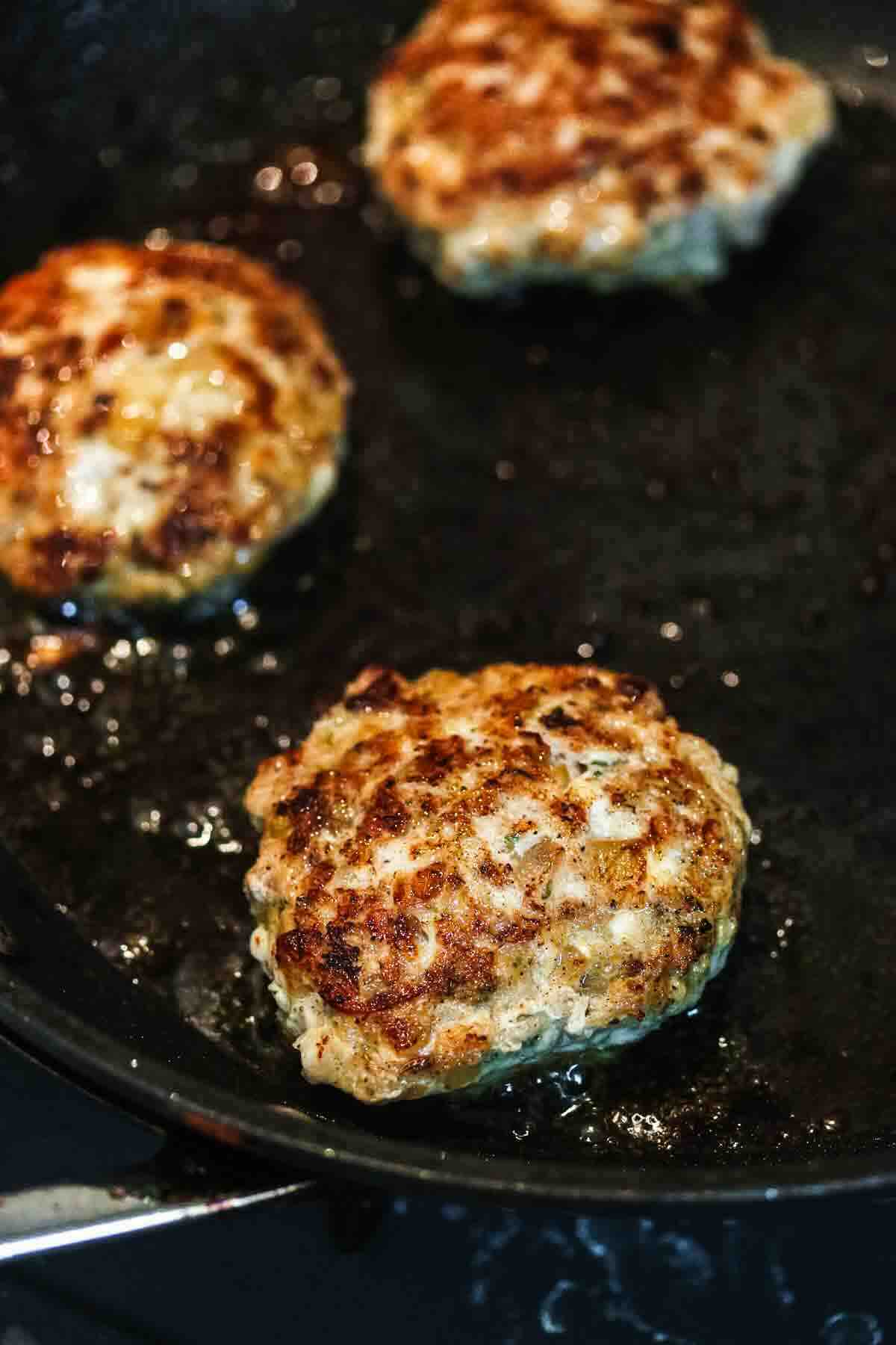 Three browned sausage patties cooking in a black skillet with visible oil.