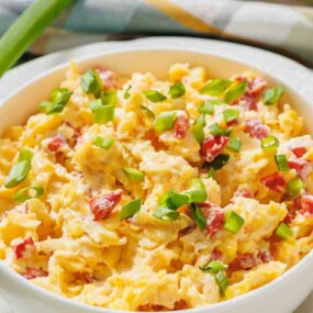 A bowl of scrambled eggs mixed with diced red peppers and topped with chopped green onions, placed on a white plate with a green onion beside it.
