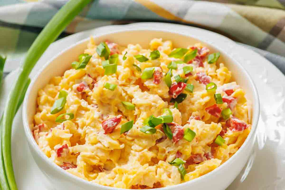 A bowl of scrambled eggs mixed with diced red peppers and topped with chopped green onions, placed on a white plate with a green onion beside it.