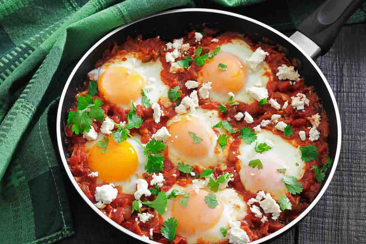 A pan of shakshuka with poached eggs in tomato sauce, garnished with fresh herbs and crumbled cheese, sits on a dark table next to a green cloth.