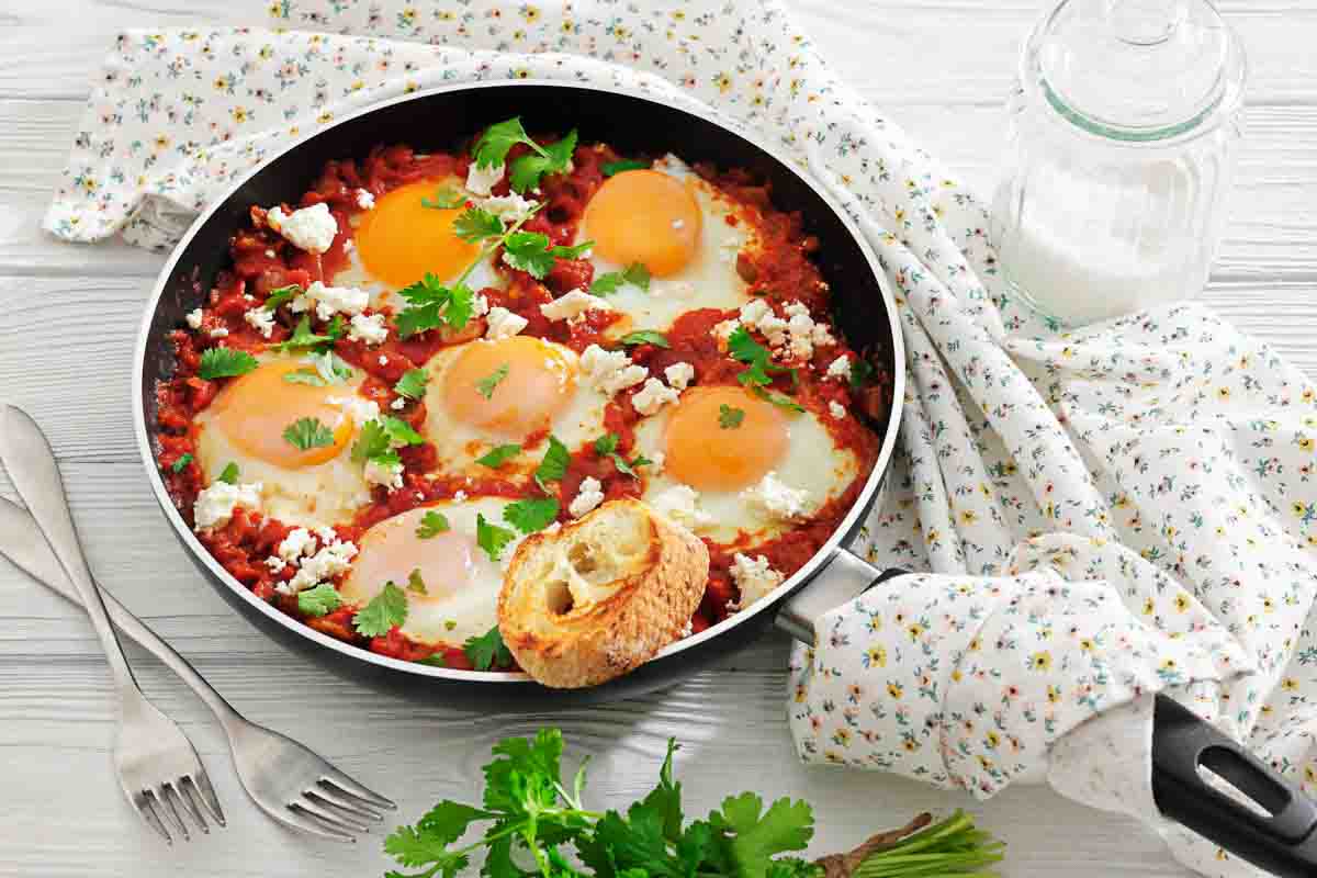 A skillet of shakshuka with poached eggs, tomato sauce, feta cheese, and parsley, served with a piece of bread. Milk, utensils, and a cloth napkin are nearby.