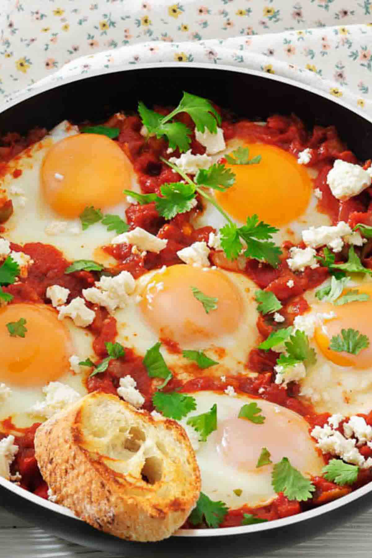 A skillet of shakshuka with six poached eggs in tomato sauce, topped with herbs and crumbled cheese, and a slice of toasted bread.