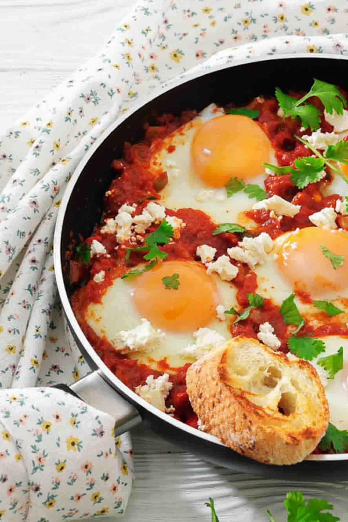 A skillet with three sunny-side-up eggs cooked in tomato sauce, garnished with parsley and feta cheese, served with a slice of toasted bread.