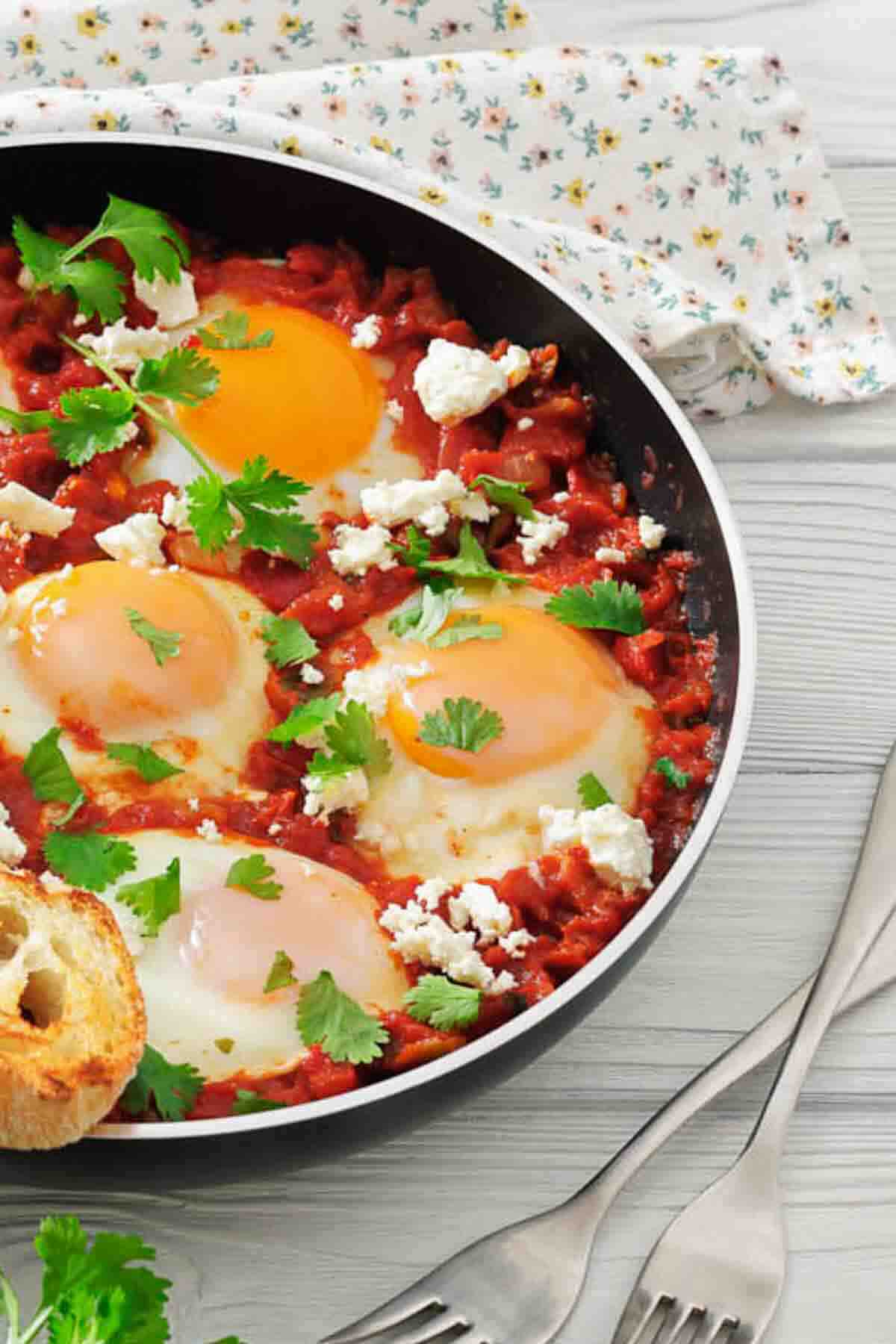 A skillet with four poached eggs in a tomato sauce, garnished with cilantro and crumbled cheese, next to a slice of bread and two forks on a table.