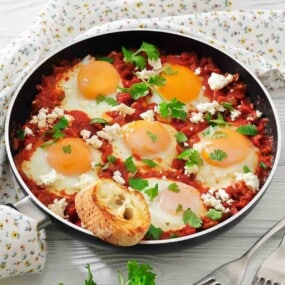 A skillet containing six eggs poached in a tomato sauce with herbs, topped with crumbled cheese and parsley, with a slice of bread on the side.