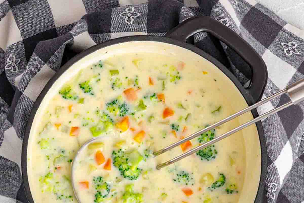 A pot of creamy broccoli and carrot soup sits on a black and white checkered cloth, with a metal ladle and whisk inside the pot.
