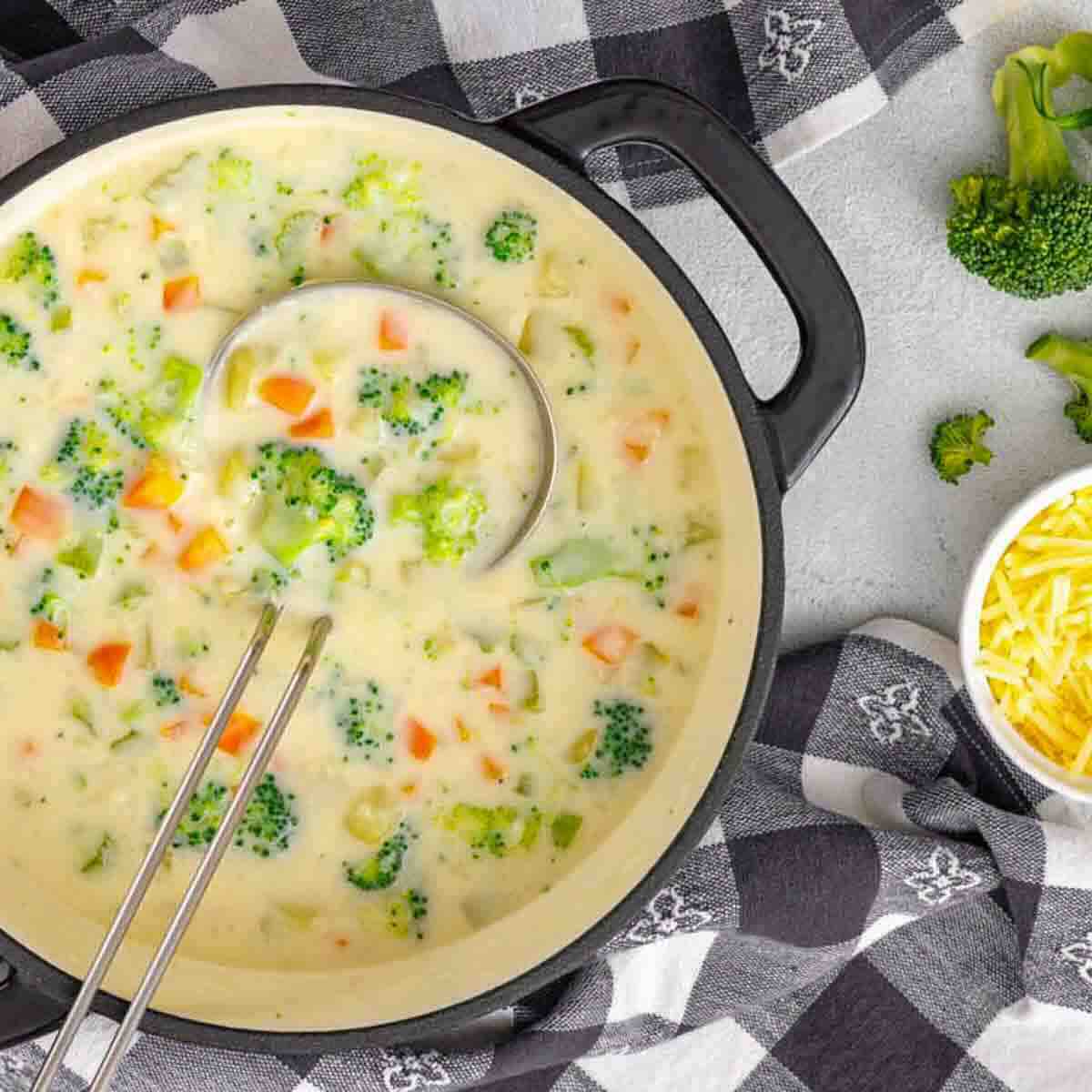 A pot of creamy broccoli and cheese soup with a ladle, surrounded by a checkered towel, shredded cheese, and broccoli florets.