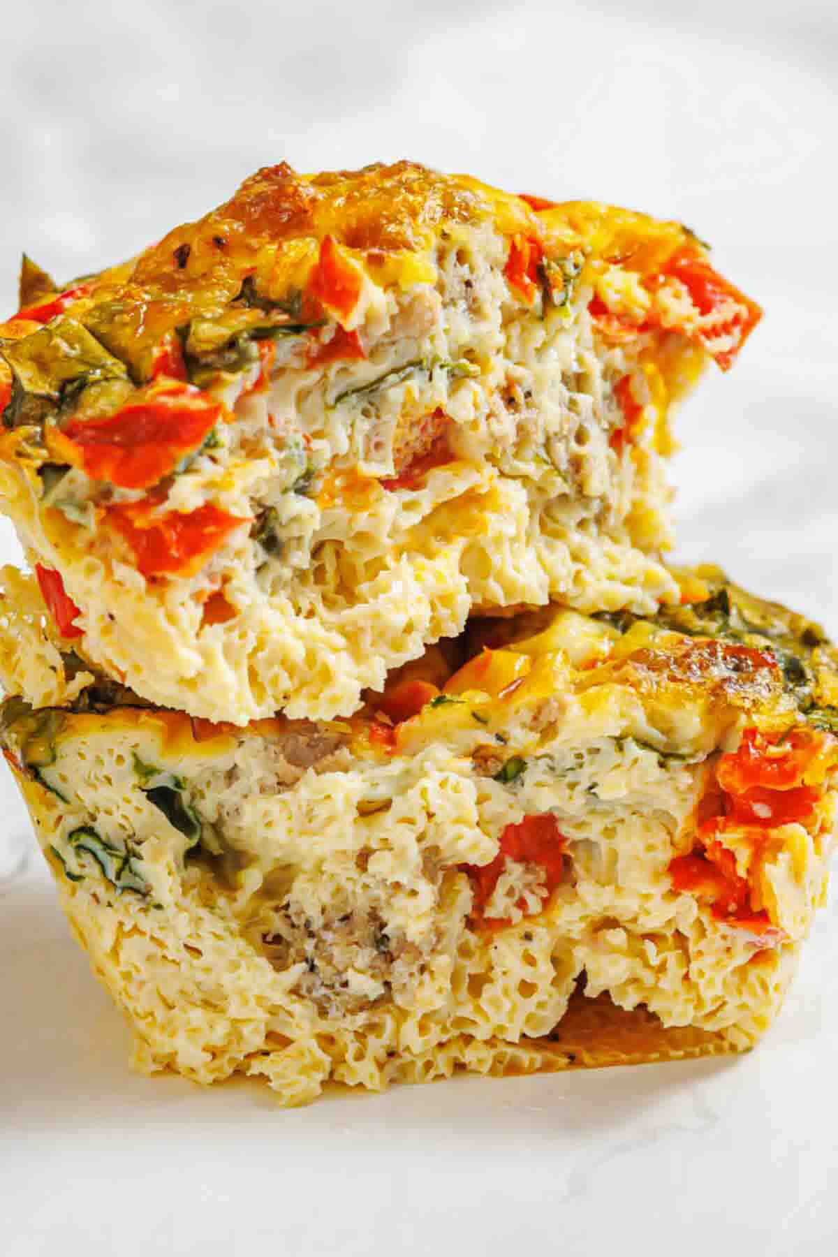 Two egg muffin cups filled with chopped vegetables, sausage, and herbs, stacked on top of each other on a light surface.