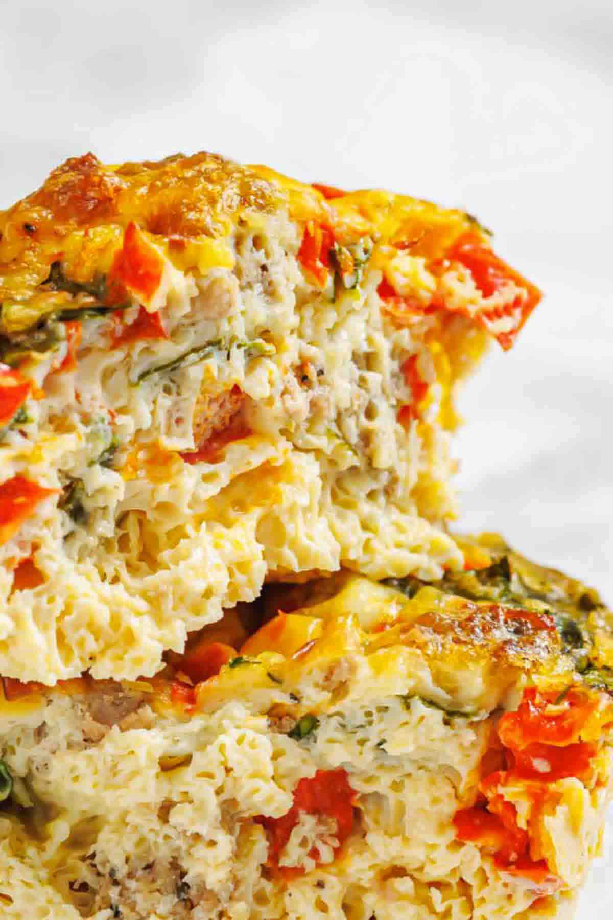 Close-up of a stacked pasta frittata with visible pieces of sausage, red bell pepper, and leafy greens, showing a creamy and textured interior.