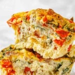 Close-up of two stacked slices of egg frittata with visible red bell peppers, greens, and melted cheese.