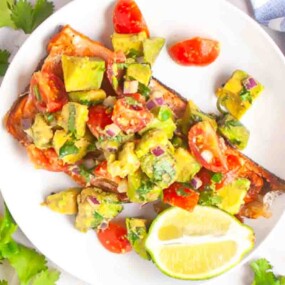 A plate with grilled salmon topped with an avocado, tomato, and onion salsa, garnished with herbs and served with a lime wedge.