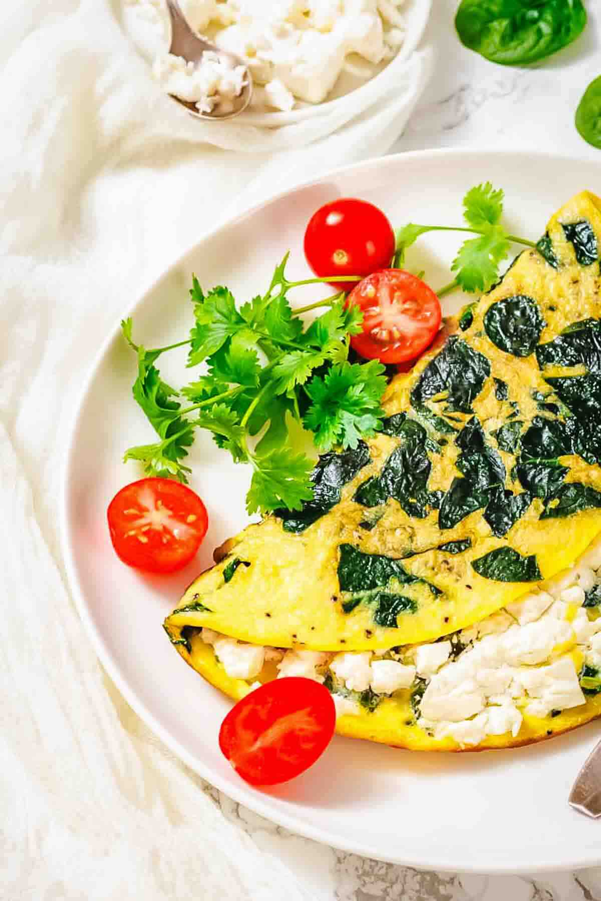 A spinach and feta omelette is served on a white plate with cherry tomato halves and a sprig of fresh cilantro, with a bowl of crumbled feta in the background.