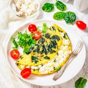 A spinach and cheese omelette served on a white plate with cherry tomatoes, fresh spinach leaves, and cilantro, with a fork on the side.