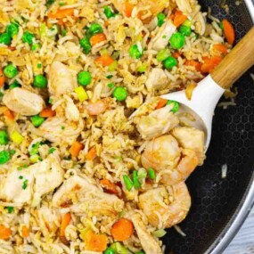 A pan filled with fried rice, chicken, shrimp, peas, carrots, and scrambled egg, with a wooden spoon scooping some of the food.