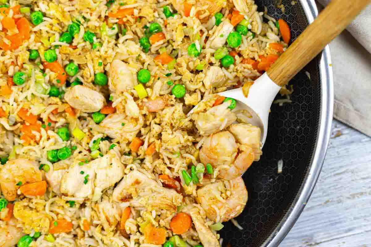 A pan filled with fried rice, chicken, shrimp, peas, carrots, and scrambled egg, with a wooden spoon scooping some of the food.
