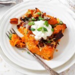 A serving of baked sweet potato casserole topped with sour cream, chopped green onions, bacon bits, and shredded cheese on a white plate with a fork.