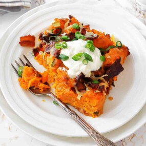 A serving of baked sweet potato casserole topped with sour cream, chopped green onions, bacon bits, and shredded cheese on a white plate with a fork.