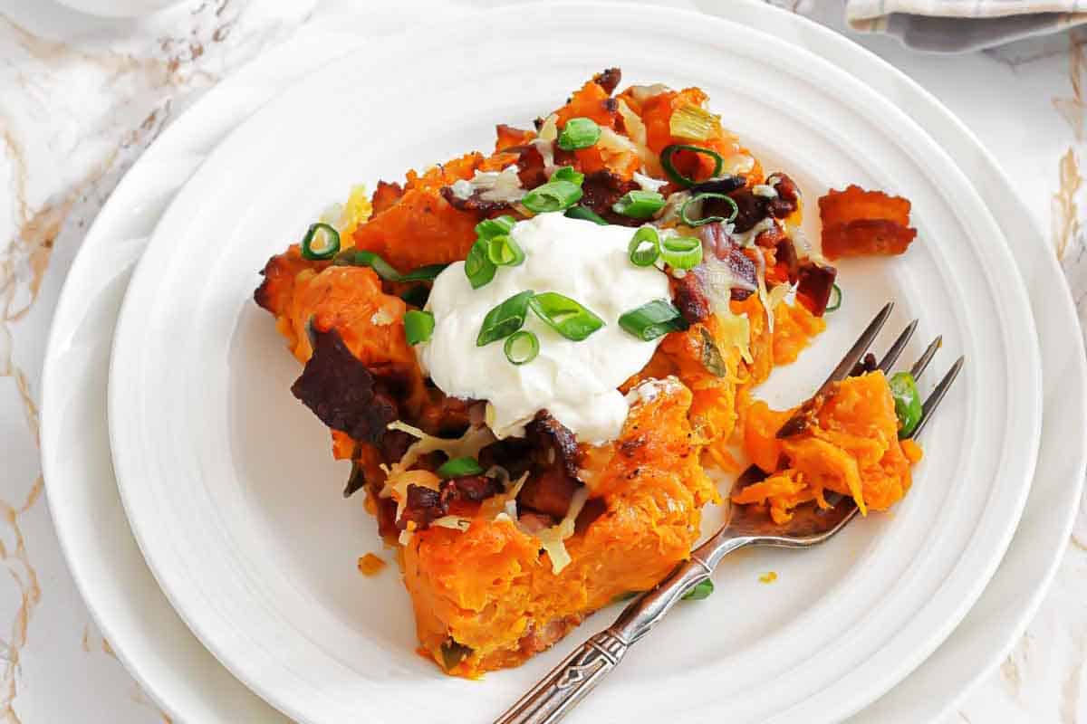 A serving of baked sweet potato casserole topped with sour cream, chopped green onions, bacon bits, and shredded cheese on a white plate with a fork.