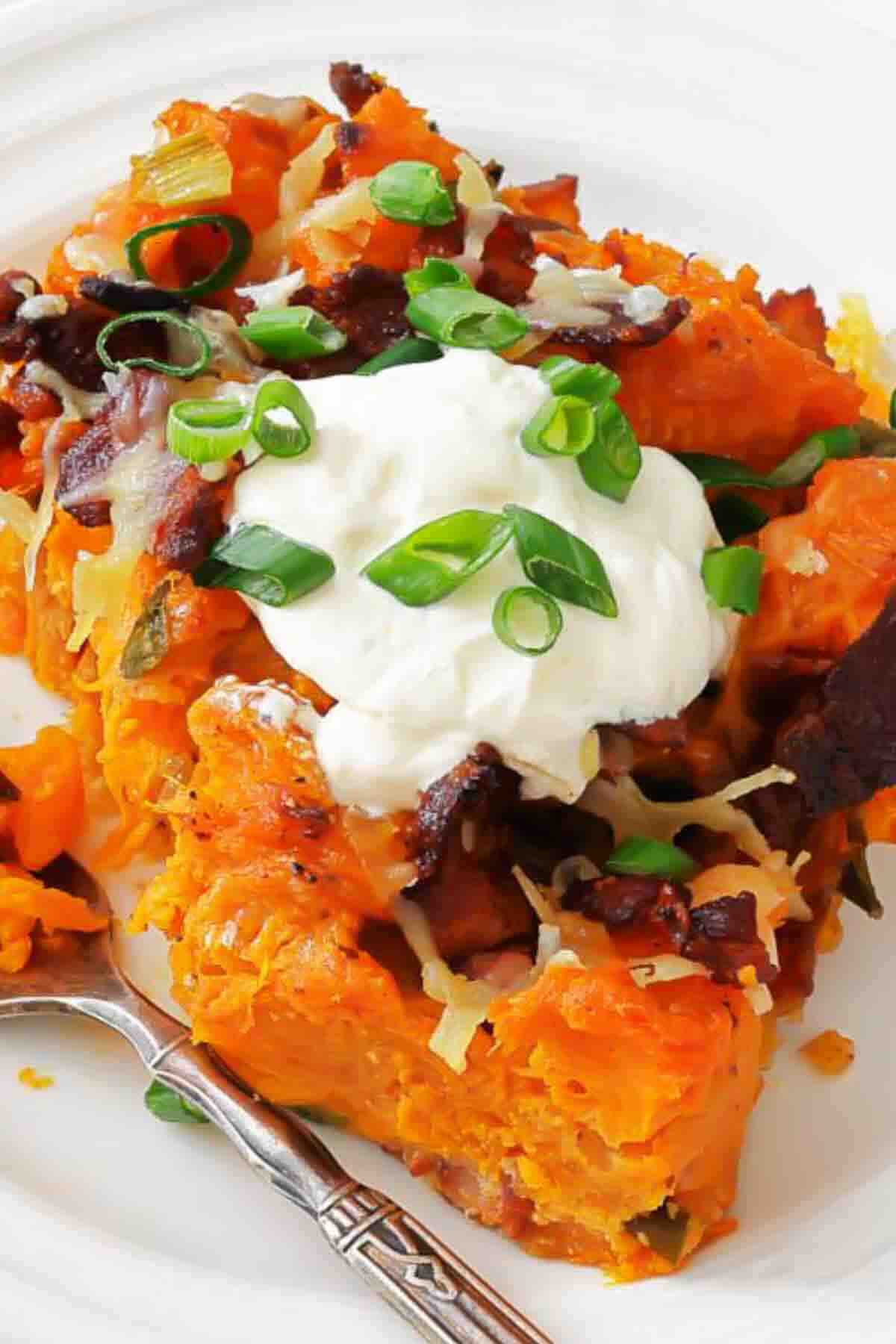 Baked sweet potato topped with sour cream, shredded cheese, bacon pieces, and chopped green onions on a white plate with a fork.