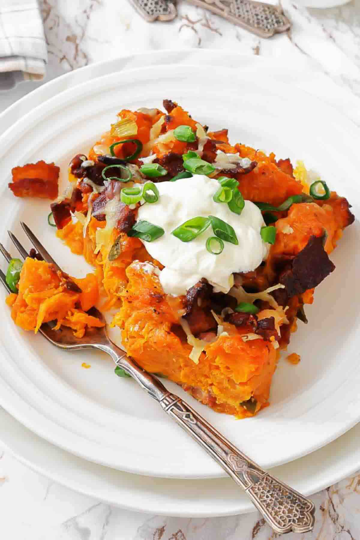 A serving of baked sweet potato casserole topped with sour cream, chopped green onions, bacon pieces, and shredded cheese on a white plate with a fork.
