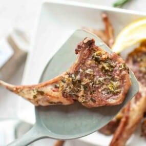 A cooked, seasoned lamb chop on a spatula, held above a plate with more chops and a lemon wedge in the background.