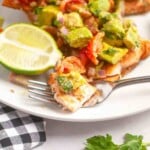 A fork holds a bite of grilled salmon topped with avocado salsa, next to a lime wedge on a white plate.