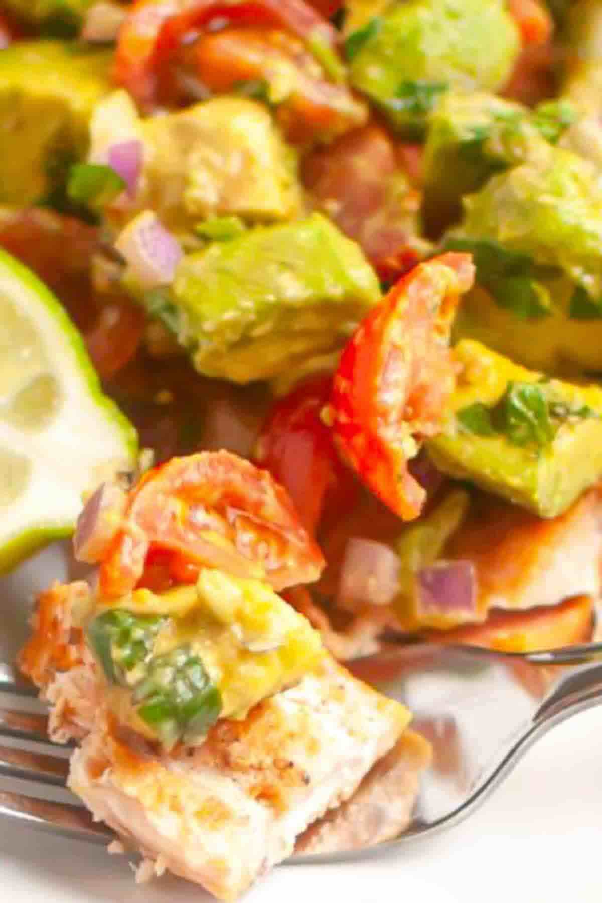 Close-up of a fork holding a piece of grilled chicken topped with avocado, tomato, red onion, and cilantro, with a slice of lime in the background.