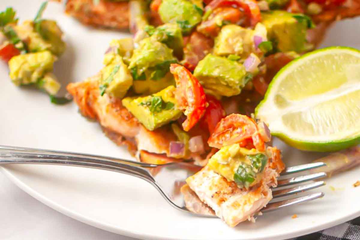 A close-up of a fork holding a bite of grilled salmon topped with avocado salsa, next to a lime wedge on a white plate.