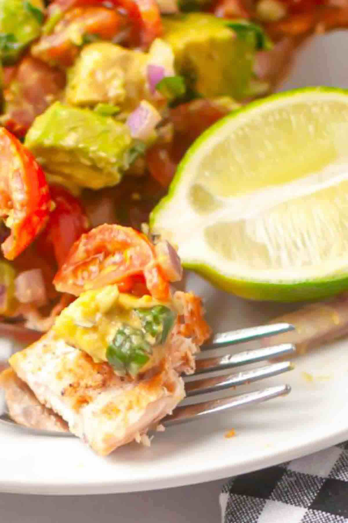A fork with a piece of grilled fish topped with avocado and tomato salsa, next to a halved lime on a white plate.